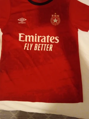 Röd ESS fotbollströja från Umbro - Etoile fotbollströja från Umbro med  sponsorerna Emirates och Ooredoo tryckta på bröstet och ryggen. Tröjan har korta ärmar, svart krage och är tillverkad i ett lätt och ventilerande material, perfekt för träning eller match.