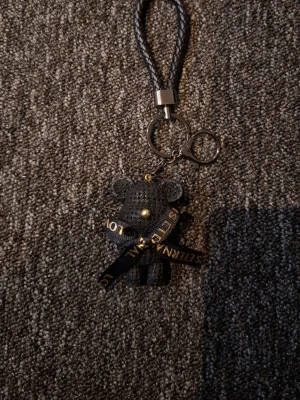 Nyckelring med svart björn och textband - Snygg nyckelring med en svart björnfigur i konstläder och ett flätat handtag. Björnen har ett dekorativt band med guldfärgad text och små gulddetaljer. Perfekt att hänga på väskan eller nycklarna för en personlig touch.