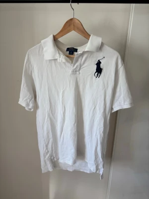 Vit pikétröja från Polo Ralph Lauren - Klassisk vit pikétröja från Polo Ralph Lauren med stor marinblå logga på bröstet och siffran 3 broderad på ärmen. Tröjan har krage, korta ärmar och knappar vid halsen. Perfekt för en clean och sportig look.
