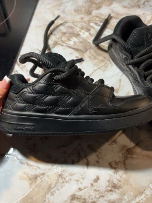 Svarta Karl Kani sneakers - Säljer ett par svarta sneakers från Karl Kani i storlek  36,5. Skorna har quiltade detaljer på sidorna, tjock sula och klassisk låg profil. Tillverkade i syntetmaterial med matchande svarta skosnören och diskret logga på sidan.