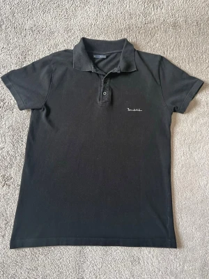 Svart pikétröja med broderad logga - Klassisk svart pikétröja med korta ärmar och krage. Tröjan har två knappar framtill och en liten vit broderad logga på bröstet. Perfekt till jeans eller shorts för en clean och enkel stil.