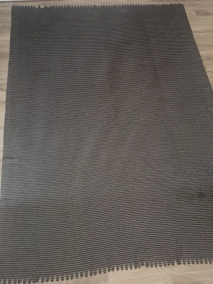 Grå filt med vita ränder - Mjuk och stor filt i grått med tunna vita ränder. Filtens struktur är ribbad och fleece tyg. Perfekt att ha i soffan eller som extra lager i sängen. Materialet känns mjukt och behagligt mot huden.
