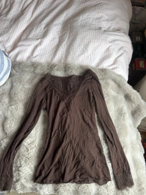 Brun långärmad topp med spetsdetaljer - Snygg brun långärmad topp med v-ringning och spetsdetaljer vid halsringning och ärmslut från intimissmi.