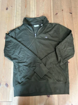 Olivgrön halvzip tröja från Gant - Olivgrön sweatshirt från Gant med halv dragkedja och ståkrage. Tröjan har broderad logga på bröstet och ribbade muddar vid ärmslut och nederkant. Perfekt för en avslappnad och stilren look.