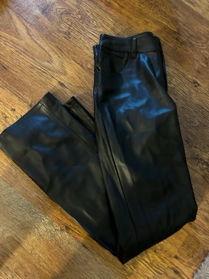 Svarta raka fuskskinnjeans - Säljer ett par svarta jeans i fuskskinn med raka ben och klassisk femficksmodell. Byxorna har bälteshällor och snygg glansig finish som ger en cool look. Perfekta för dig som vill sticka ut lite extra.