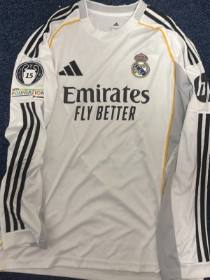 Real Madrid långärmad matchtröja - Säljer en vit långärmad Real Madrid matchtröja från Adidas med svarta detaljer och gula inslag. Tröjan har klubbmärke, sponsortryck och Champions League-märke på ärmen. Namn och nummer 'Rodrygo 11' på ryggen. Perfekt för fotbollsfans!