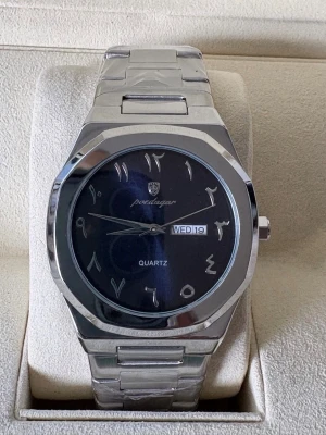 Silverfärgad Poedagar klocka med blå urtavla - Stilren klocka från Poedagar med silverfärgat armband i metall och blå urtavla. Urtavlan har arabiska siffror och dag/datumvisning. Klassisk design som passar till många stilar.