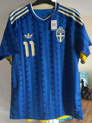 Blå Sverige fotbollströja Elanga 11 - Säljer en blå Sverige fotbollströja från Adidas med Elanga och nummer 11 på ryggen. Tröjan har gula detaljer, klassiska Adidas-ränder på axlarna och ett mönster i blå nyanser. Perfekt för dig som vill stötta landslaget med stil.