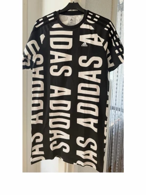 Svart kort klänning med vit Adidas-logga - Cool svart kort sports klänning från Adidas med stora vita loggor tryckta över hela klänning. Klassisk rund hals och korta ärmar. Perfekt för dig som gillar sportig och trendig streetstyle. Materialet är mjukt och bekvämt.