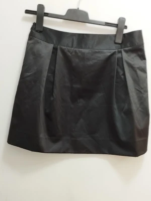 Svart kortkjol i satin - Elegant svart kortkjol med enkel design och diskreta veck framtill. Kjolen har en rak passform och är tillverkad i ett mjukt satin som känns mjukt och inte blir skrynkligt. Perfekt för en både vardag och uppklätt! För mått se bilderna. Kolla gärna mina andra annonser <3
