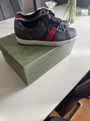Gucci Ace sneakers svart/röd/blå - Gucci Ace sneakers i svart med klassiskt GG-mönster, röd och blå rand på sidan samt metallicröd och metallicblå detalj på hälen. Låg modell med vita sulor och svarta skosnören. Lyxig känsla och ikonisk design i läder och textil.