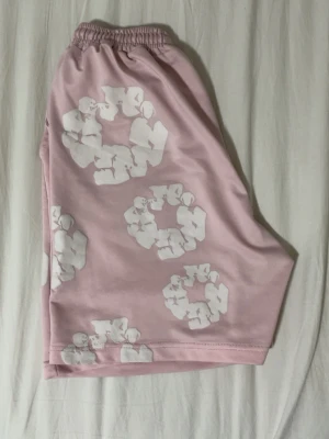 Denim ters rosa shorts - Säljer ett par ljusrosa shorts med elastisk midja och vita grafiska mönster över hela plagget. Shortsen har en loose passform och är perfekta för sommaren. Materialet känns mjukt och svalt, troligen polyester eller bomull.