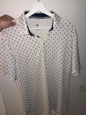 Vit Golfpiké med blått mönster från Adidas - Vit pikétröja från Adidas med små blåa trianglar över hela tyget. Klassisk krage och knappar framtill. Perfekt för en sportig och avslappnad stil. Stor i storleken 