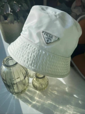 Vit bucket hat från Prada - Pris kan diskuteras Snygg vit bucket hat från Prada i glansigt nylonmaterial. Klassisk design med det ikoniska triangulära Prada-emblemet framtill. Perfekt accessoar för en trendig och avslappnad stil.  Box ingår 