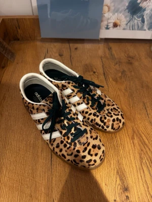 Adidas sneakers med leopardmönster - Unika Adidas sneakers med leopardmönstrad ovandel i mocka och klassiska vita ränder på sidorna. Skorna har mörkgröna skosnören, vit insida och brun gummisula. Perfekta för dig som vill sticka ut med din stil.