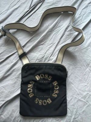 Svart axelväska från BOSS - Snygg svart axelväska från BOSS med justerbar beige axelrem och logga i guld på framsidan. Väskan har en fyrkantig form och dragkedja upptill. Perfekt för att bära det viktigaste och ger en clean look till din outfit.