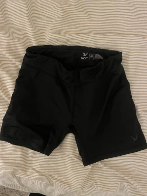 Svarta tighta träningsshorts SOC - Svarta träningsshorts från SOC med tight passform och midwaist. Tillverkade i ett stretchigt syntetmaterial som passar perfekt för gymmet eller löpning. Klassisk design utan synliga detaljer förutom loggan på benet. Dem passar även dem som är xxs/xs