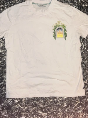 Vit Casablanca t-shirt - Säljer en vit t-shirt med tryck från Casablanca. T-shirten har ett färgglatt motiv med ett fönster och gröna blad både fram och bak. Klassisk passform och rund halsringning. Perfekt för dig som gillar unika prints.