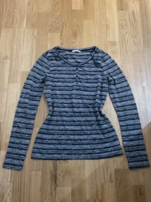 Gråmelerad långärmad topp - Snygg gråmelerad långärmad topp med smala svarta och grå ränder. Rund halsringning och knappdetaljer framtill. Figurnära passform som framhäver midjan. Perfekt att matcha med jeans eller kjol för en avslappnad look.