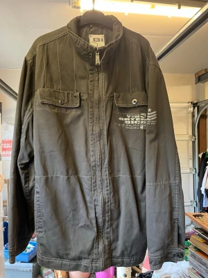 Olivgrön jacka från Bison - Olivgrön overshirt från Bison i slitstark bomull. Jackan har två bröstfickor med lock och knapp, dragkedja framtill och tryckdetalj på vänster bröst. Ståkrage och raka sömmar ger en clean och avslappnad look. Perfekt för lager på lager och streetwear-vibe. Axel till axel ca:50cm. Axel till midjan ca:68. Arm ca:60cm. Midja ca:55cm. 