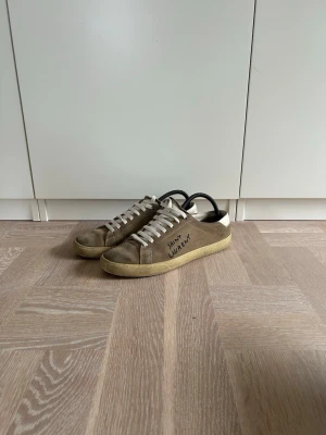 Saint Laurent bruna låga sneakers - Snygga bruna sneakers från Saint Laurent med vit sula och vita skosnören. Skorna har en klassisk låg siluett och är tillverkade i mocka med diskret logga på sidan. Perfekta för en avslappnad och trendig stil.