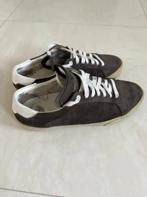 Saint Laurent gråa låga sneakers - Gråa sneakers från Saint Laurent i mocka med vita detaljer och vita skosnören. Skorna har vit hälkappa med logotyp och beige sula. Klassisk och stilren design som passar till många olika outfits.