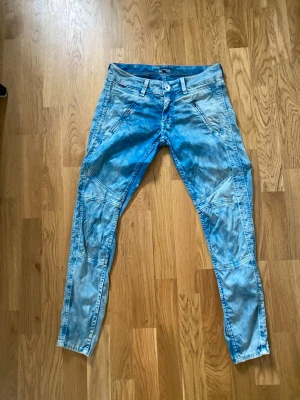 Tommy Hilfiger Denim – Sophie Biker 7/8 Jeans – W27/L32 (S) - Supercoola vintage bikerjeans från Hilfiger Denim (modellen Sophie Biker 7/8). Syratvättad blå nyans med tuffa mc-detaljer över knäna och dragkedjor. Sitter tight och supersnyggt med låg midja (low waist). Från okt 2013.Material: 97% Lyocell, 3% Elastan (mycket skön stretch!)Storlek: W27/L32 (ca 36/S)Midja: 38 cm, Innerben: 68 cm, Gren: 19 cmSkick: Gott skick, dragkedjor funkar. En söm har släppt lite på ena knät (se bild).