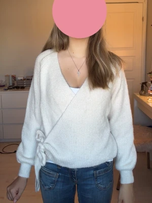 Vit omlottstickad tröja - Mysig vit stickad omlott-tröja med knytband i sidan och ribbade muddar. Tröjan har en djup V-ringning och ballongärmar som ger en trendig look. Perfekt att matcha med jeans för en avslappnad stil.