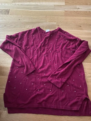 Vinröd tröja med pärlor från Zara - Vinröd långärmad tröja från Zara Girls med små vita pärlor som dekoration framtill. Tröjan har rund halsringning och är tillverkad i ett mjukt, tunt material som känns skönt mot huden. Perfekt för dig som gillar en enkel men ändå snygg stil.
