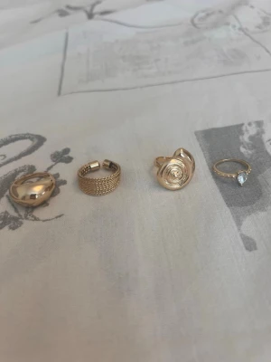 Fyra guldfärgade ringar i olika stilar - Säljer ett set med fyra guldfärgade ringar i olika designer. En har en chunky, rundad form, en har flätad meshstruktur, en har en spiralformad dekoration och en är tunn med en klar sten. Perfekta för att mixa och matcha till din stil.