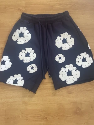 Mörkblåa shorts med vita blommor - Säljer ett par mörkblåa shorts med stora vita blommönster och råa kanter. Shortsen har resårmidja med snörning för skön passform och är perfekta för en avslappnad stil.