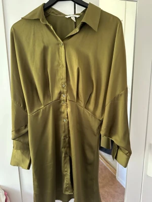 Olivgrön skjortklänning från Zara - Snygg olivgrön skjortklänning från Zara i glansigt material. Klänningen har knappar framtill, krage och långa ärmar med lös passform. Perfekt för dig som gillar stilrena och trendiga plagg med en avslappnad vibe.