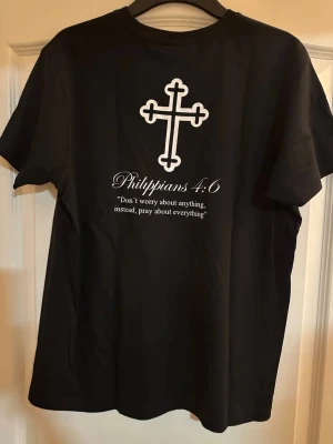 Svart t-shirt med kors och citat - Svart t-shirt med vitt kors på bröstet och större kors med texten 'Philippians 4:6' samt citat på ryggen. Klassisk passform och rund halsringning. Perfekt för dig som vill ha en stilren och uttrycksfull look.