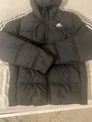 Svart dunjacka med huva från Adidas - Snygg svart dunjacka från Adidas med huva och dragkedja framtill. Jackan har de klassiska vita Adidas-ränderna längs axlarna och logga på bröstet. Puffig modell som håller dig varm under kalla dagar. Perfekt för vintern och riktigt street.