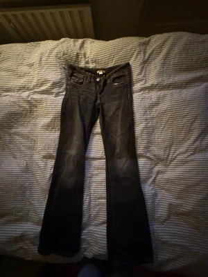 Svarta bootcut jeans med slitningar - Säljer ett par svarta bootcut jeans med snygga slitningar framtill och klassiska fem fickor. Jeansen har en låg midja och silverfärgade knappar. Perfekta för dig som gillar en avslappnad men trendig stil.