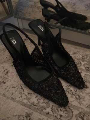 Svarta paljettpumps från Zara - Säljer ett par svarta pumps från Zara med spetsig tå och öppen häl. Skorna är täckta med glittrande paljetter och har en elegant slingback-rem. Inte använda och i storlek 36.