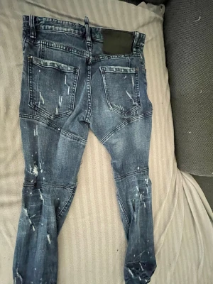 Blå Dsquared2 jeans med slitningar - Säljer ett par blå jeans från Dsquared2 i storlek 44. Jeansen har coola slitningar och färgstänk för en edgy look, samt markerade sömmar över benen och bakfickor. Tillverkade i klassiskt denimtyg som ger en snygg passform.