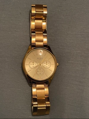Guldfärgad klocka från Regal - Stilren guldfärgad klocka från Regal med metallarmband och rund urtavla. Klockan har tre dekorativa subdials och minimalistiska markeringar. Perfekt accessoar för dig som gillar en klassisk och elegant look.