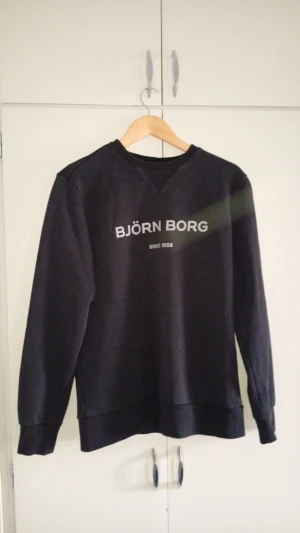Svart sweatshirt från Björn Borg - Klassisk svart sweatshirt från Björn Borg med tryckt logga och texten 'Since 1956' på bröstet. Tröjan har rund halsringning, ribbade muddar vid ärmslut och nederkant samt en relaxed passform. Perfekt till jeans eller joggers.