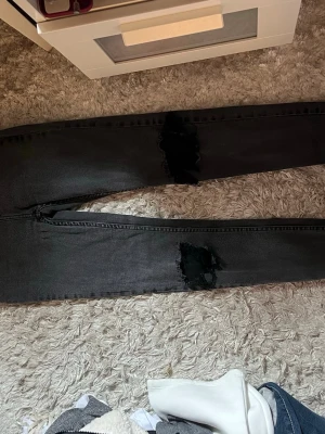 Svarta ripped skinny jeans - Säljer ett par svarta jeans från lager157 med stora slitningar över knäna. Jeansen har en smal passform och klassisk femficksdesign. Byxorna är högmidjade och Perfekta för dig som gillar en edgy och trendig stil.