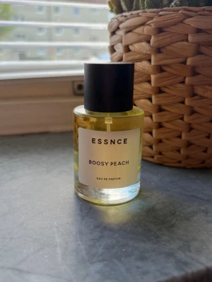 ESSNCE Boosy Peach Eau de Parfum - En stilren parfymflaska från ESSNCE med doften Boosy Peach. Flaskan är cylindrisk i genomskinligt glas med svart rund kork och vit etikett. Innehållet är ljusgult och ger ett fräscht och modernt intryck. Perfekt för dig som gillar fruktiga dofter. Ny 