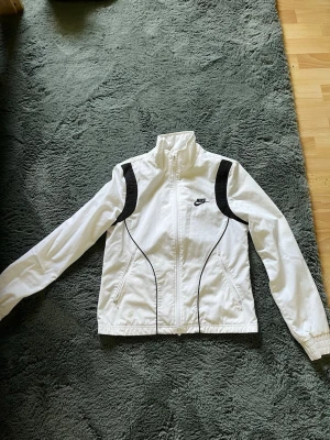  Nike jacka - Snygg vit träningsjacka från Nike med svarta detaljer på axlar och längs sidorna. Jackan har dragkedja framtill, hög krage och två fickor med dragkedja. Perfekt för sport eller chill dagar. Tillverkad i lätt och bekvämt material.
