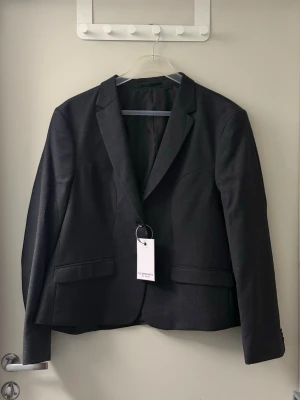 Svart kavaj med klassisk passform - Stilren svart kavaj med klassisk passform och två fickor framtill. Kavajen har smala slag och stängs med en knapp. Perfekt för en dressad look och enkel att matcha med olika outfits. Materialet ser ut att vara en blandning med slät yta.