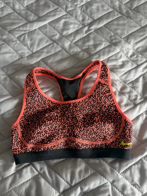 Nike sport-bh med neonmönster - Snygg sport-bh från Nike med svart och neonkorallfärgat abstrakt mönster. Brottarrygg och elastisk svart kant nedtill. Liten neongul Nike-logga framtill. Perfekt för träning och sportiga aktiviteter.