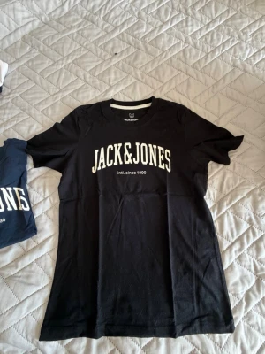 Svart Jack & Jones t-shirt - Svart t-shirt från Jack & Jones med vit logga tryckt på bröstet. Klassisk rund halsringning och korta ärmar. Perfekt för en avslappnad och stilren look.
