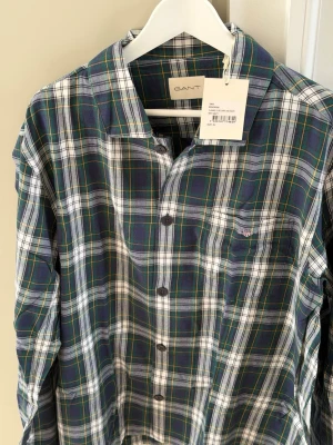 Grönblå rutig skjorta från GANT - Pyjamasskjorta från GANT i grönt, blått och vitt rutmönster med tunna gula linjer. Skjortan har klassisk krage, knäppning framtill och är tillverkad i flannel. Aldrig använd med prislapp kvar. 