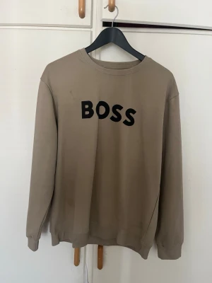 Beige sweatshirt från BOSS - Säljer en beige sweatshirt från BOSS med svart tryck på bröstet. Tröjan har rund halsringning, ribbade muddar vid ärmslut och nederkant samt en avslappnad passform. Perfekt för en casual och trendig look.