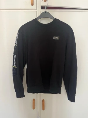 Svart sweatshirt från EA7 Emporio Armani - Snygg svart sweatshirt från EA7 Emporio Armani med tryckt logga på bröstet och detaljer längs ena ärmen. Tröjan har rund halsringning, ribbade muddar och en avslappnad passform. Perfekt för en sportig och trendig look.