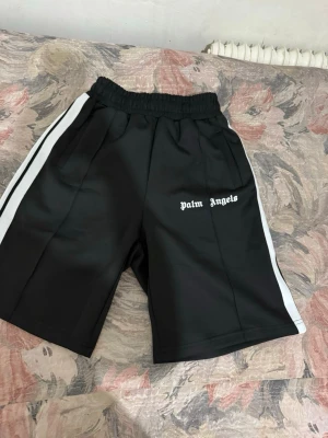 Svarta shorts med vita ränder Palm Angels - Säljer ett par svarta shorts från Palm Angels med vita sidoränder och logga framtill. De har elastisk midja och en loose passform, perfekta för en avslappnad streetwear-look. Materialet känns som polyester och shortsen är knälånga.