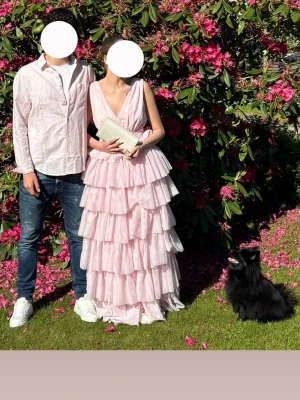 Ljusrosa långklänning med volanger - Säljer en ljusrosa långklänning med flera lager volanger och djup v-ringning. Klänningen är ärmlös och har en omlott-topp samt öppen rygg. Perfekt för sommarens speciella tillfällen. Bara andvänd en gång.